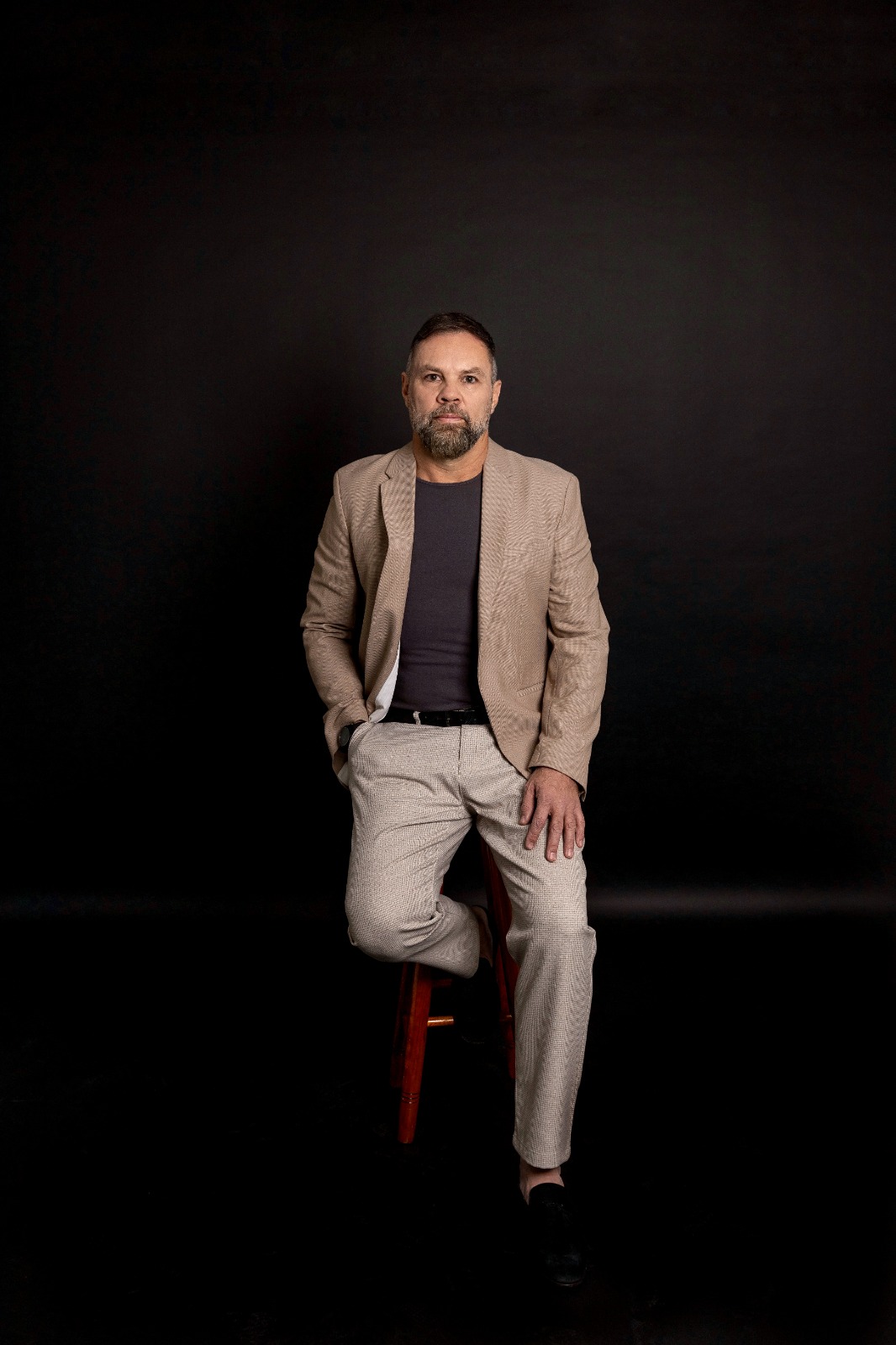 Retrato editorial de Paulo Tavares sentado, de blazer bege e camiseta escura